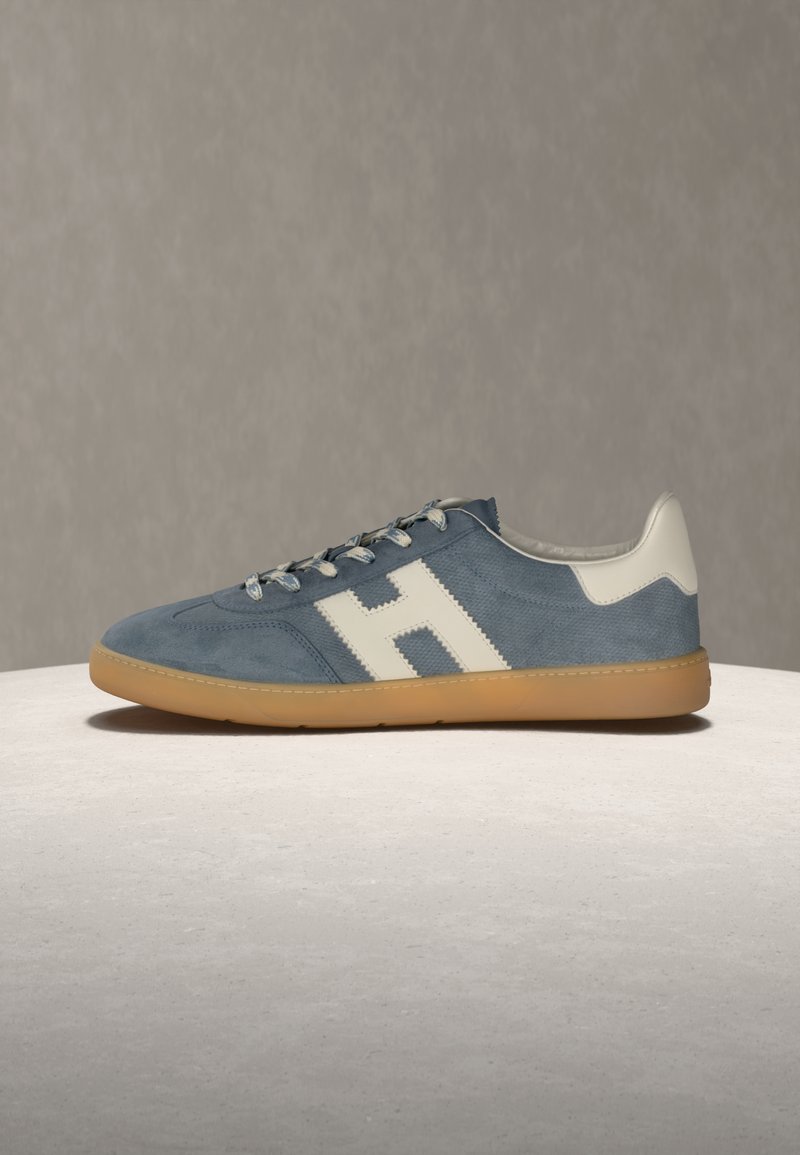 Μπλε σουέντ αθλητικό παπούτσι με λευκό γράμμα "H" στο πλάι, λευκό ταμπελάκι στη φτέρνα, γκρι κορδόνια και σόλα από καουτσούκ χρώματος καραμέλας σε ανοιχτή επιφάνεια.