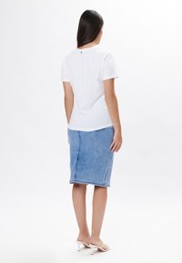 Camiseta blanca de manga corta con cuello redondo, combinada con una falda lápiz de mezclilla azul claro y sandalias de tacón blancas. Vista desde atrás.