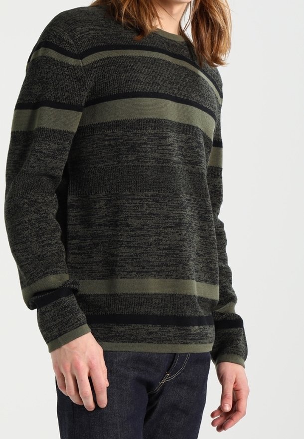 Personne portant un pull à rayures gris foncé et vert olive à manches longues et un jean bleu foncé, debout devant un fond clair uni.