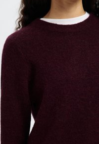 Bordo pleten pulover z okroglim izrezom, teksturirano tkanino in dolgimi rokavi. Nošen preko belega topa, ima mehak, puhast zaključek.