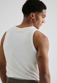 Hvid ribbet tanktop med rund halsudskæring. Den tætsiddende design fremhæver skuldre og arme. Har en yin-yang tatovering på højre arm.