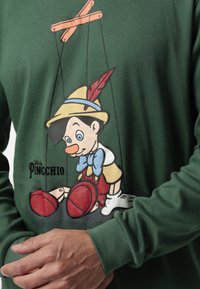 Camisa de manga larga verde con una ilustración a color de Pinocho jugando con cuerdas de marioneta. Los detalles incluyen unos zapatos rojos y un sombrero con pluma.