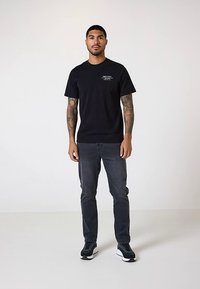 Hombre con brazos tatuados lleva una camiseta gráfica negra, jeans oscuros y zapatillas negras, de pie contra un fondo claro y liso.