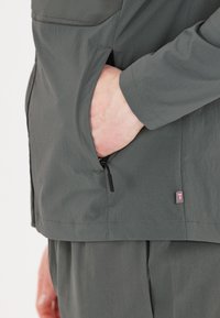 Grå teknisk jacka med dragkedjefickor och en liten logootag. Tyget har en slät yta med ett lätt utseende.