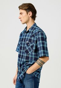 Camicia a maniche corte a quadri con tonalità blu e navy, due tasche sul petto e un design con bottoni e colletto classico.