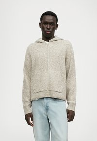 Μπεζ πλεκτή ζακέτα με φερμουάρ στο μπροστινό μέρος, μεγάλες μπροστινές τσέπες και κουκούλα με κορδόνι. Η υφή είναι ραβδωτή, με μια λεπτή πολυτονική σχέδια.