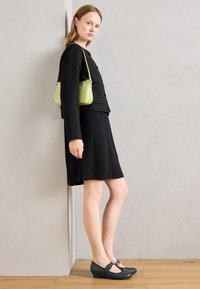 Woman in black skirt, black jacket, black low-heeled shoes, and light green shoulder bag, standing sideways against a light gray wall.