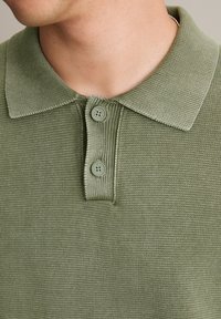 Zielona dzianinowa koszulka polo z teksturowanej tkaniny, z dwoma guzikami i prążkowanym kołnierzykiem, nadająca zorganizowany wygląd.