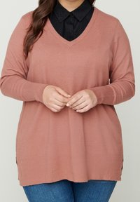 Maglione rosa chiaro a scollo a V con maniche lunghe, polsini a coste e spacchi laterali. Indossato sopra una camicia nera con colletto decorata con borchie.