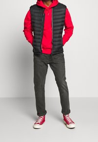 Un gilet trapuntato nero sopra un hoodie rosso, jeans grigi e scarpe alte rosse. Il gilet presenta una cerniera e una costruzione leggera.