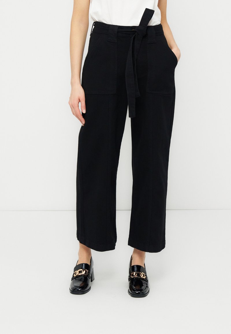 Γυναίκα που φοράει μαύρο παντελόνι με φαρδιά μπατζάκια, μπροστινές τσέπες και δεμένη ζώνη, συνδυασμένο με μαύρα παπούτσια λουστρίνι με χρυσή αλυσίδα.