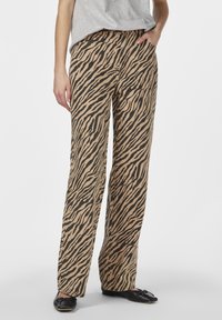 Beige bukser med et sort tigerstribemønster, der har lige ben og sidelommer, parret med sorte flade sko.