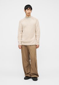 Jeune homme portant un pull beige texturé, un pantalon large marron et des chaussures noires, debout devant un fond clair uni.