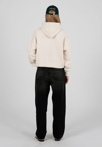 Felpa con cappuccio color crema con una grande tasca posteriore, abbinata a jeans neri a vita alta. Il modello indossa un berretto verde scuro.