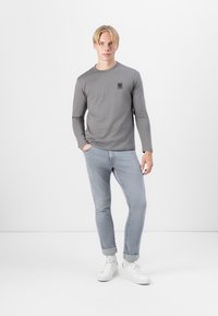 Cămașă gri cu mânecă lungă și guler rotund, cu un mic patch logo negru, asociată cu blugi slim-fit albastru deschis și adidași albi.