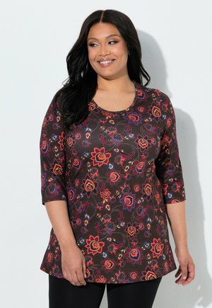 Kvinde med langt mørkt hår iført en brun top med blomsterprint og trekvartærmer samt sorte bukser, smilende og kiggende til siden.