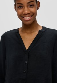Haut noir à boutons avec un col en V et un tissu texturé. Présente une finition douce et trois boutons sur le devant pour la fermeture.