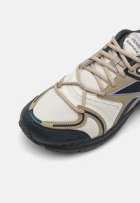 Shoe atlético leve com parte superior em malha nas cores creme e areia, sola em borracha preta, ressaltos laterais em azul e atacadores texturizados.