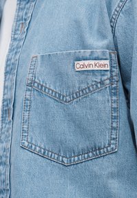 Τσέπη πουκάμισου από ανοιχτό μπλε τζιν με καφέ ραφές, που φέρει μια λευκή ετικέτα με κόκκινο κείμενο "Calvin Klein" στο πάνω δεξιό μέρος.