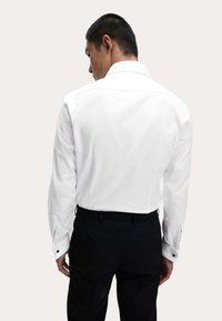 Mann steht mit dem Rücken zur Kamera, trägt ein weißes, langärmeliges Hemd mit Kragen, das in eine schwarze Hose gesteckt ist, vor einem schlichten Hintergrund.