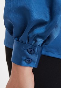 Blaues Satinhemd mit gerafftem Design an den Manschetten, versehen mit zwei dunkelblauen Knöpfen zum Schließen. Der Stoff hat eine dezente gerippte Textur.
