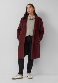 Frau, die gegen eine schlichte Wand steht und einen kastanienbraunen Mantel, einen grauen Schalkragenpullover, schwarze Hosen und weiße Sneaker mit beigen Sohlen trägt.