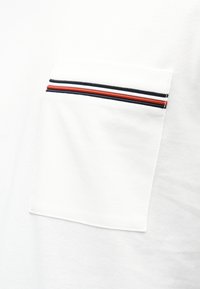 Λευκό πόλο μπλουζάκι με τσεπάκι στο στήθος που έχει οριζόντιες ρίγες σε κόκκινο, λευκό και ναυτικό μπλε. Η υφή είναι ομαλή με ένα διακριτικό σχέδιο.