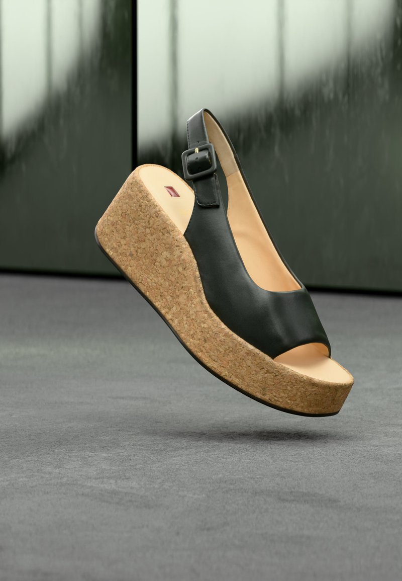 Μαύρο δερμάτινο πέδιλο με ανοιχτά δάχτυλα, πλατφόρμα φελλού και λουράκι στον αστράγαλο που αιωρείται πάνω από γκρι δάπεδο και πράσινο θολωμένο φόντο.