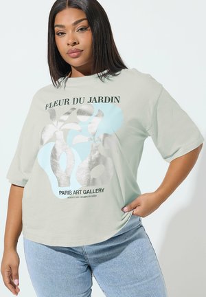 Młoda kobieta ubrana w jasnoszarą, oversize'ową koszulkę z srebrno-niebieskim abstrakcyjnym motywem roślinnym i napisem "Fleur du Jardin Paris Art Gallery", w połączeniu z niebieskimi dżinsami.
