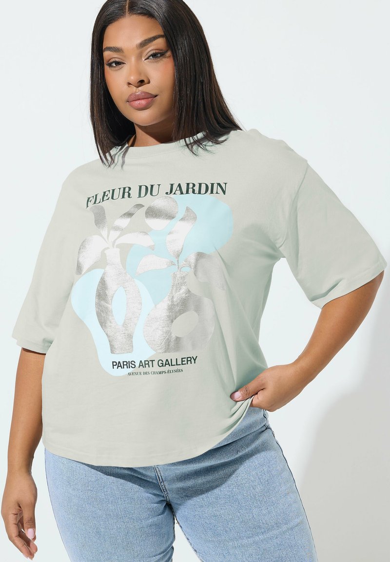 Jeune femme portant un t-shirt ample gris clair avec un motif abstrait de plante argenté et bleu et le texte "Fleur du Jardin Paris Art Gallery", associé à un jean bleu.