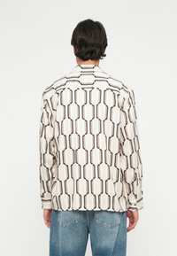 Camicetta a maniche lunghe in tessuto crema chiaro con motivo geometrico nero. Include un colletto a punta e dettagli sui polsini con bottoni.