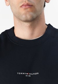Sweatshirt azul-marinho com decote redondo. Apresenta um logotipo branco "TOMMY HILFIGER" e o texto "EST. 1985" ao centro, design minimalista.