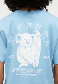 Πίσω μέρος γαλάζιου πουκάμισου που δείχνει έναν λευκό κοάλα που κρατάει μια σανίδα σερφ με το κείμενο "Rip Curl Australia Ultimate Surfing Company".