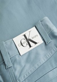 Tessuto di jeans azzurro chiaro con una texture liscia. Presenta un'etichetta bianca con "Calvin Klein Jeans" in lettere nere in grassetto. Dettagli di cucitura visibili.