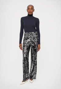 Mulher com um polo preto, calças largas de lantejoulas prateadas e pretas e sapatos de salto branco com bico fino, de pé contra um fundo simples.