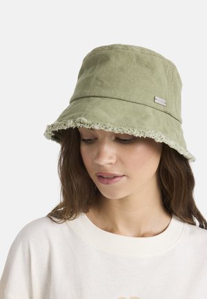 Jonge vrouw met een lichtgroene bucket hat met rafelige randen en een wit overhemd, kijkt naar beneden tegen een effen achtergrond.