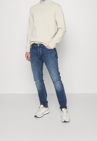 Svetle béžová mikina, modré džíny s odřením a poškozením na kolenou, doplněné béžovými a bílými teniskami. Stojící pozice.