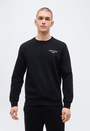 Mlajši moški s kratkimi lasmi, oblečen v črno majico Björn Borg in ujemajoče se črne hlače, stoji pred enobarvnim svetlim ozadjem.