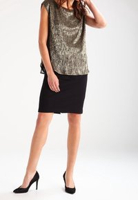 Top corto metálico dorado brillante combinado con una falda negra ajustada; el modelo lleva tacones altos negros. Textura suave y ajuste relajado.