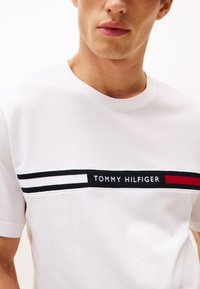 Fiatal férfi, fehér Tommy Hilfiger pólót visel, amelyen fekete, fehér és piros vízszintes logósáv fut a mellkasán.