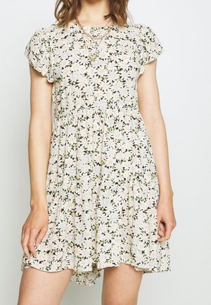 Femme portant une robe à manches courtes à motif floral avec des colliers en or superposés, posant les mains détendues le long du corps.