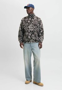 Fleece-pullover med høj krave i mørkebruin med et hvidt geometrisk mønster. Kombineret med lyseblå bukser med vidde og brune støvler.