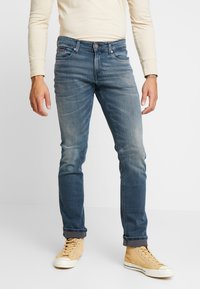 Man som bär blekta blå jeans med upprullade benslut, beige högskor och en långärmad krämfärgad skjorta, stående på vit golv.