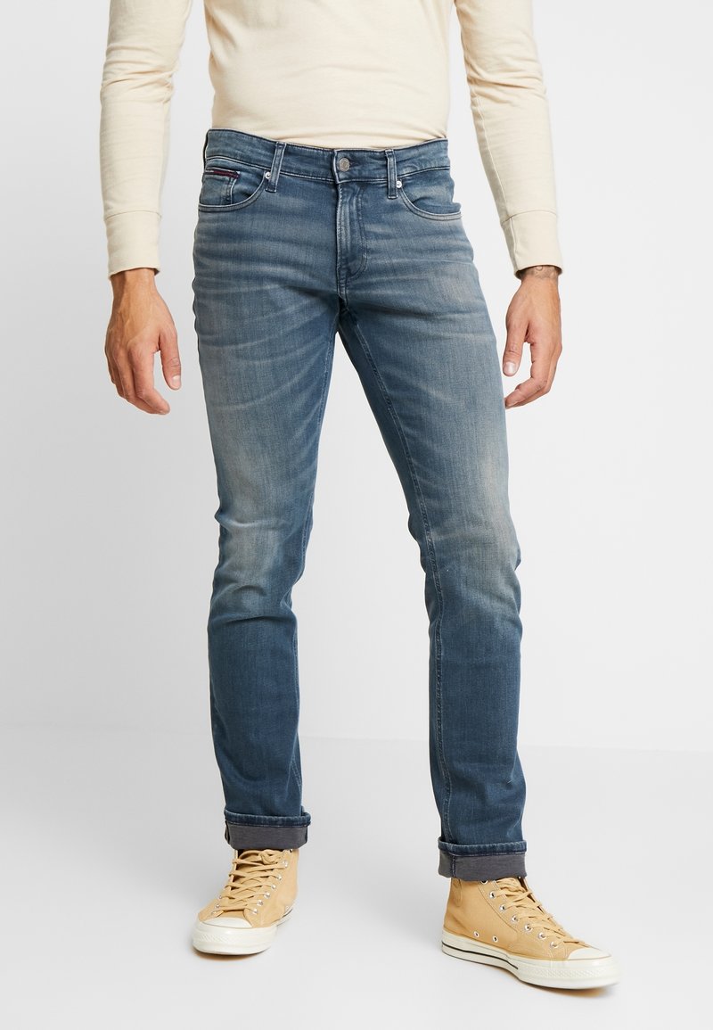 Man som bär blekta blå jeans med upprullade benslut, beige högskor och en långärmad krämfärgad skjorta, stående på vit golv.
