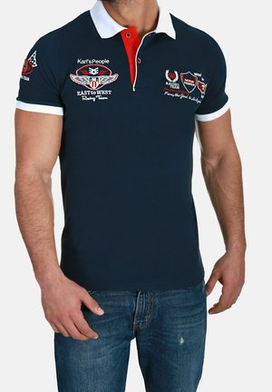 Man die een donkerblauw poloshirt met witte kraag en raceteam-logo's draagt, gecombineerd met een blauwe spijkerbroek, staat met ontspannen armen.