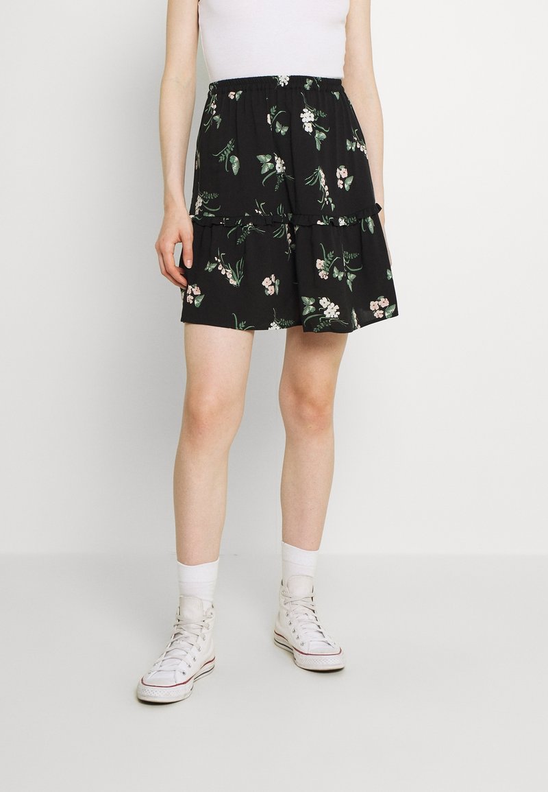 Personne portant une mini-jupe à volants noire à motifs floraux, un débardeur blanc, des chaussettes blanches et des baskets blanches montantes sur un fond uni.