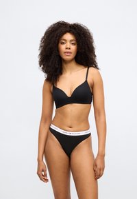 Črni modrček z globokim izrezom in tankimi trakovi, skupaj s črnimi tangicami z belim pasom, na katerem je napis "TOMMY HILFIGER".