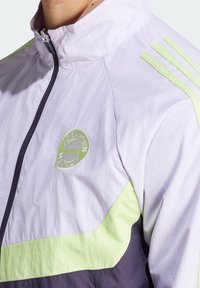 Chaqueta de color lila claro con acentos en verde neón y azul marino, con un alto cuello y el logo del FC Bayern de Múnich. Fabricada con un material suave y ligero.