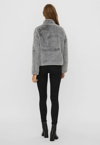 Šedá umělá kožichová bunda s vysokým límcem a dlouhými rukávy, zkrácený design, nošena přes černé úzké džíny, model otočený zády.