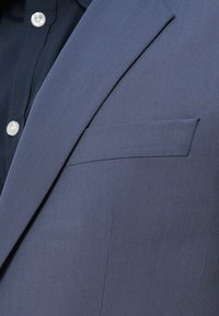 Ναυτικό μπλε σακάκι με πέτο, υφαντό ύφασμα και οριζόντια τσέπη εισιτηρίων, συνδυασμένο με μαύρο πουκάμισο με κουμπιά.